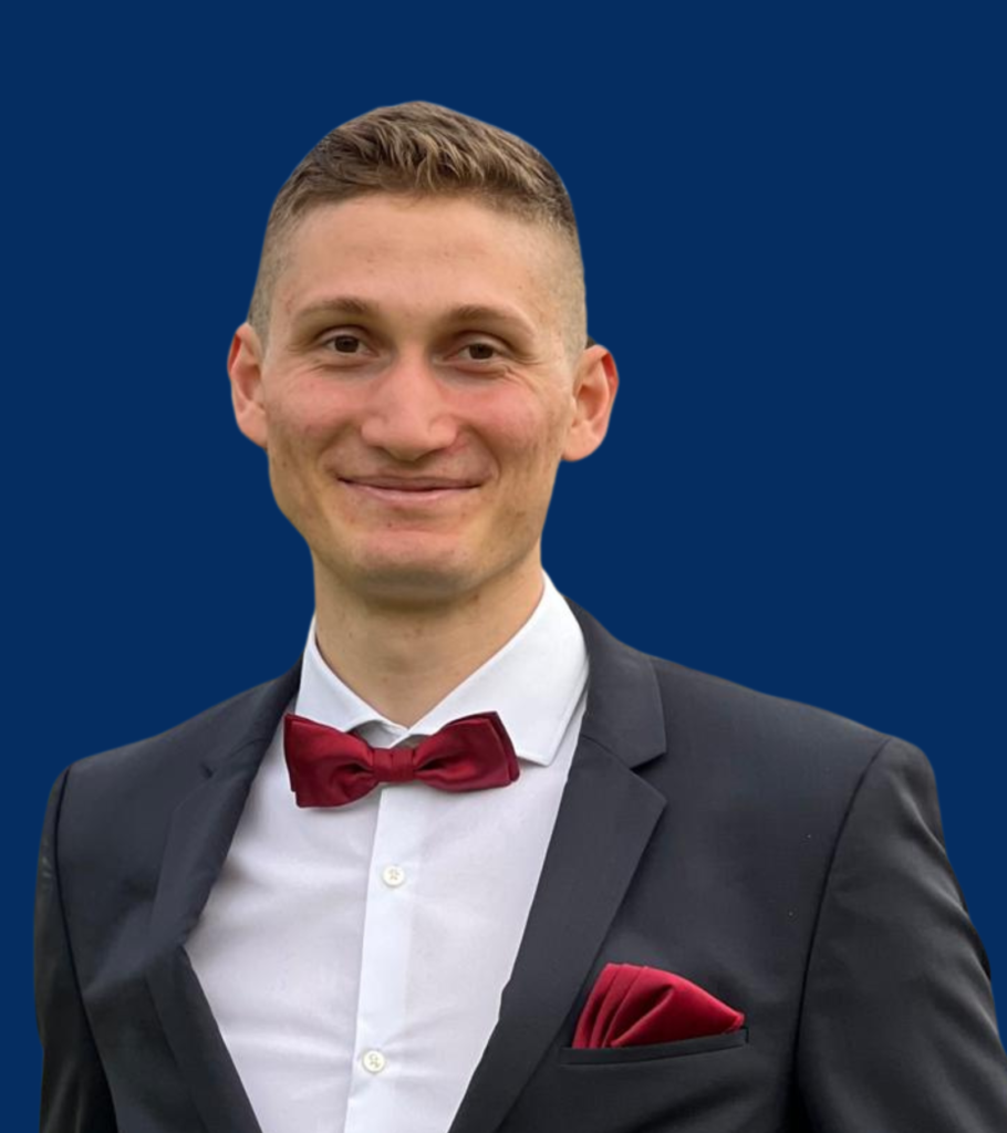 A blond boy with short hair, wearing a black suit with a red bow tie and white shirt.