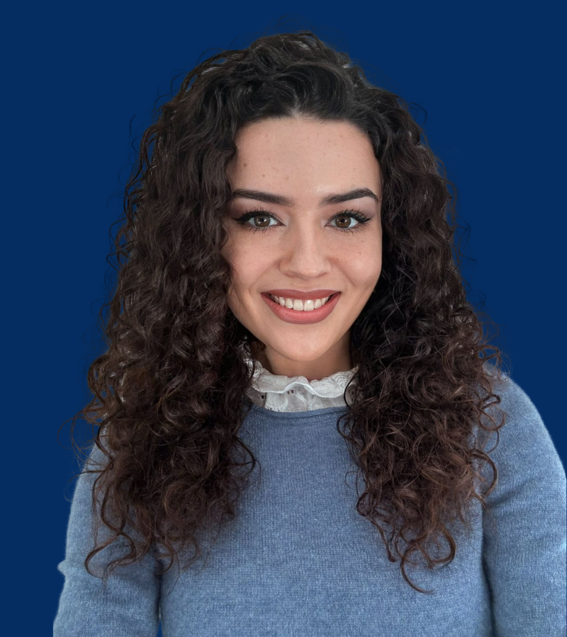 A headshot of a smiling girl with dark, long and curly hair, wearing a light blue blouse, smiling at the camera.