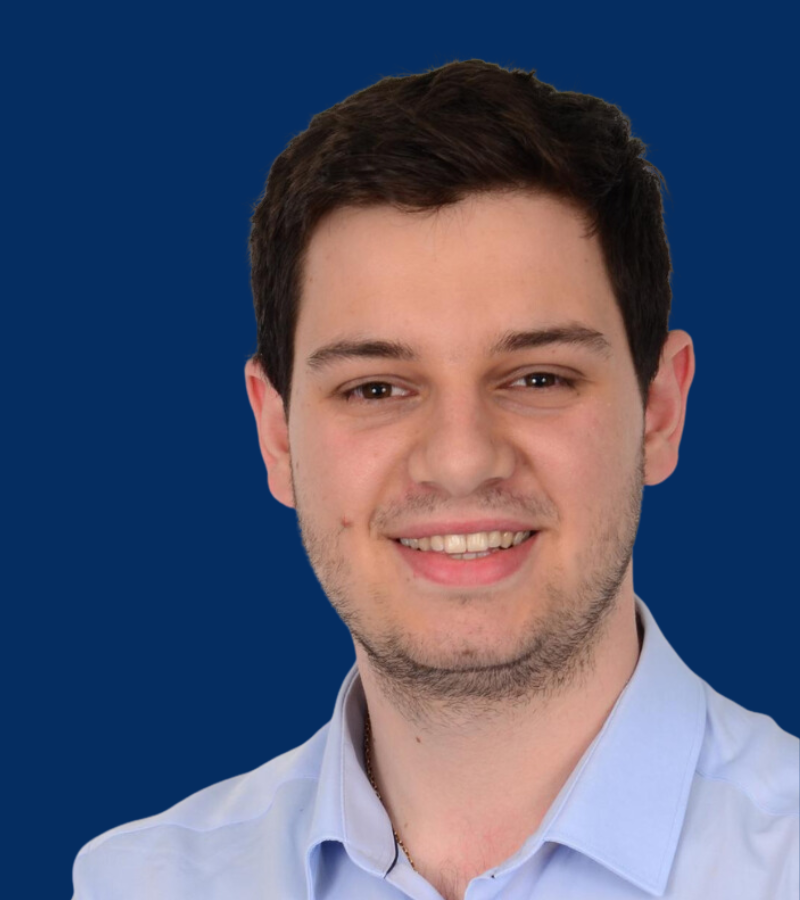 A headshot of a boy with short brown hair, wearing a light blue shirt, smiling at the camera.